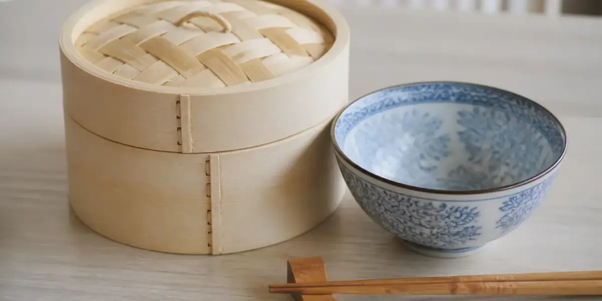 Panier vapeur en bambou fermé posé à côté d’un bol bleu et blanc sur une table claire