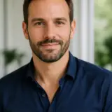 Portrait de Bastien Lorrain, cheveux bruns courts, barbe courte, chemise bleu marine, fond de pièce lumineuse et floue