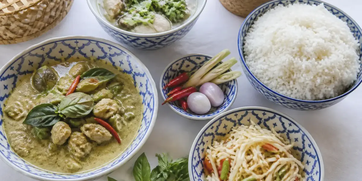 Plusieurs plats thaïlandais partagés autour d’un grand bol de riz, avec herbes fraîches et piments
