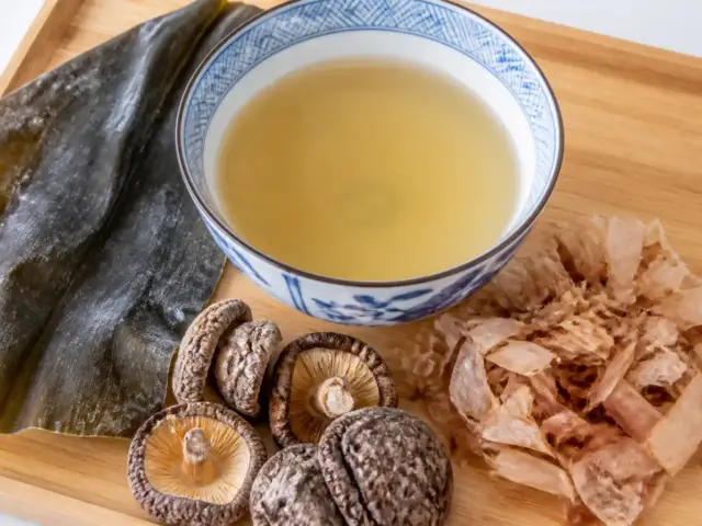 Bol de bouillon clair entouré d’algue kombu séchée, de champignons shiitake et de copeaux de bonite