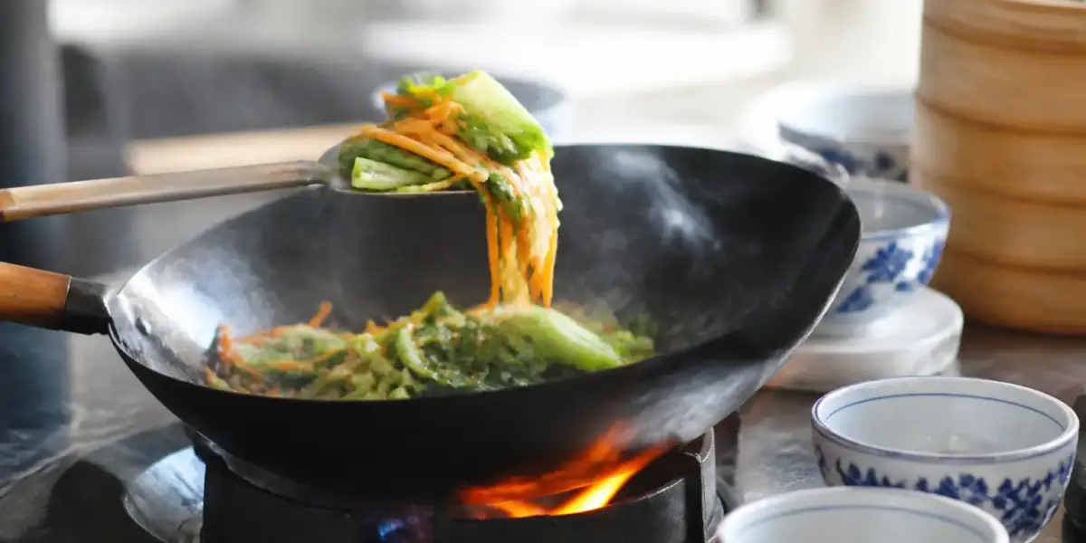 Légumes coupés en lamelles qui sautent dans un wok noir au-dessus d’une flamme vive, spatule en métal
