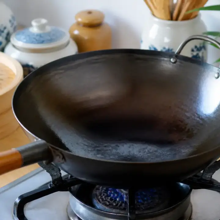 Wok en acier noirci posé sur une flamme, sa paroi intérieure luisante d’une fine couche d’huile
