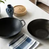 Un wok en fonte épais et un wok en acier noir plus fin posés côte à côte sur un plan de travail clair