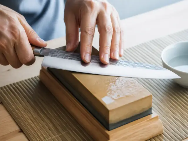 Lame de couteau japonais inclinée sur une pierre à aiguiser mouillée posée sur un socle en bois