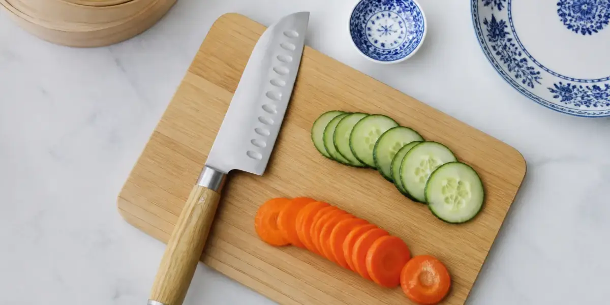 Couteau santoku à lame alvéolée posé sur une planche en bois à côté de légumes émincés