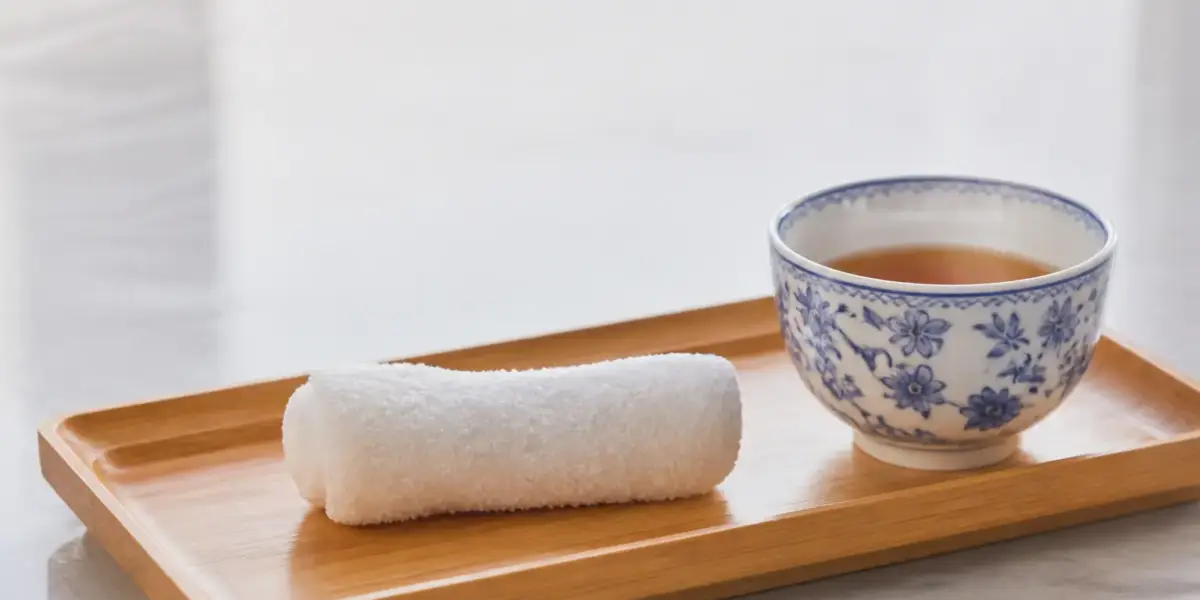 Serviette humide roulée posée sur un petit plateau en bambou, à côté d’une tasse de thé en porcelaine