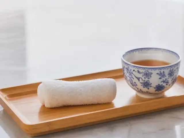 Serviette humide roulée posée sur un petit plateau en bambou, à côté d’une tasse de thé en porcelaine